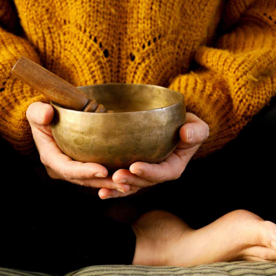 Une femme est assise en tailleur. Elle porte un pull chaud en maille orange et un pantalon noir. Elle tient dans ses main un bol tibétain en bronze et un maillet en bois.