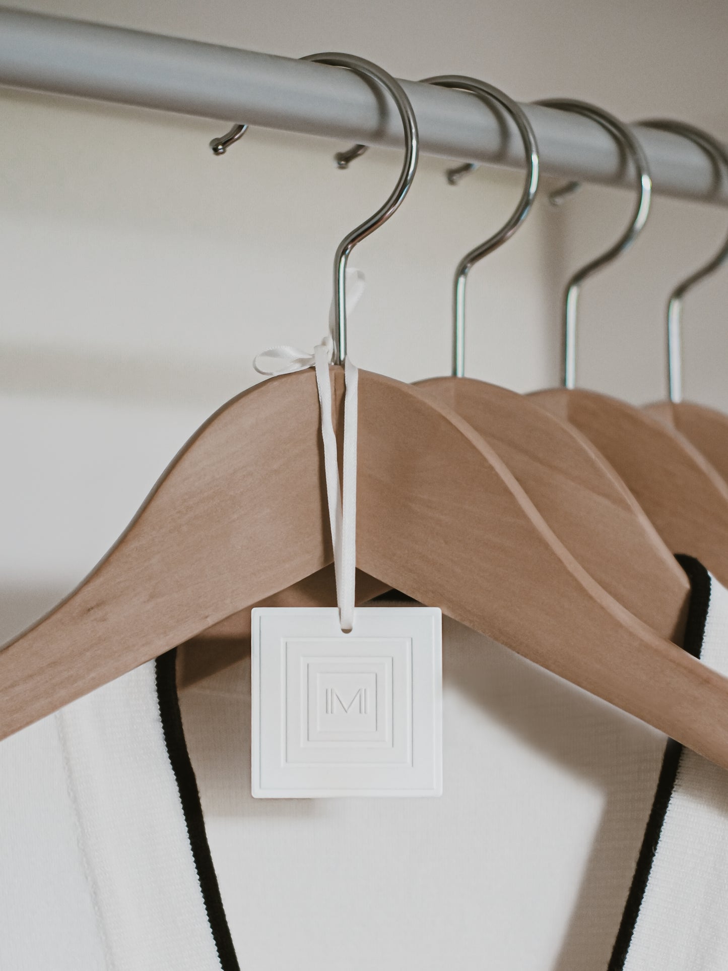 Une céramique carrée pour diffuser le parfum, blanche avec le logo gravé de la marque Marylise Mirabelli est suspendue avec son ruban satin de couleur crème à un cintre en bois dans un dressing.