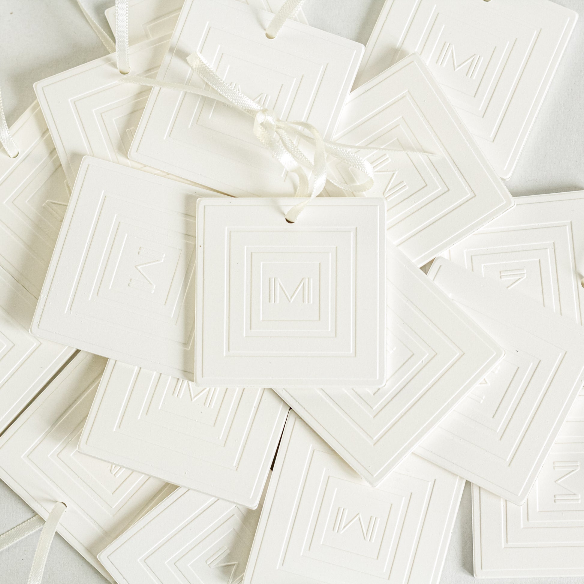 Plusieurs céramiques à parfum blanches avec le logo gravé de la marque Marylise Mirabelli sont disposées en tas. Ce sont des diffuseur d'intérieur en céramique avec leur ruban satin crème pour suspendre.