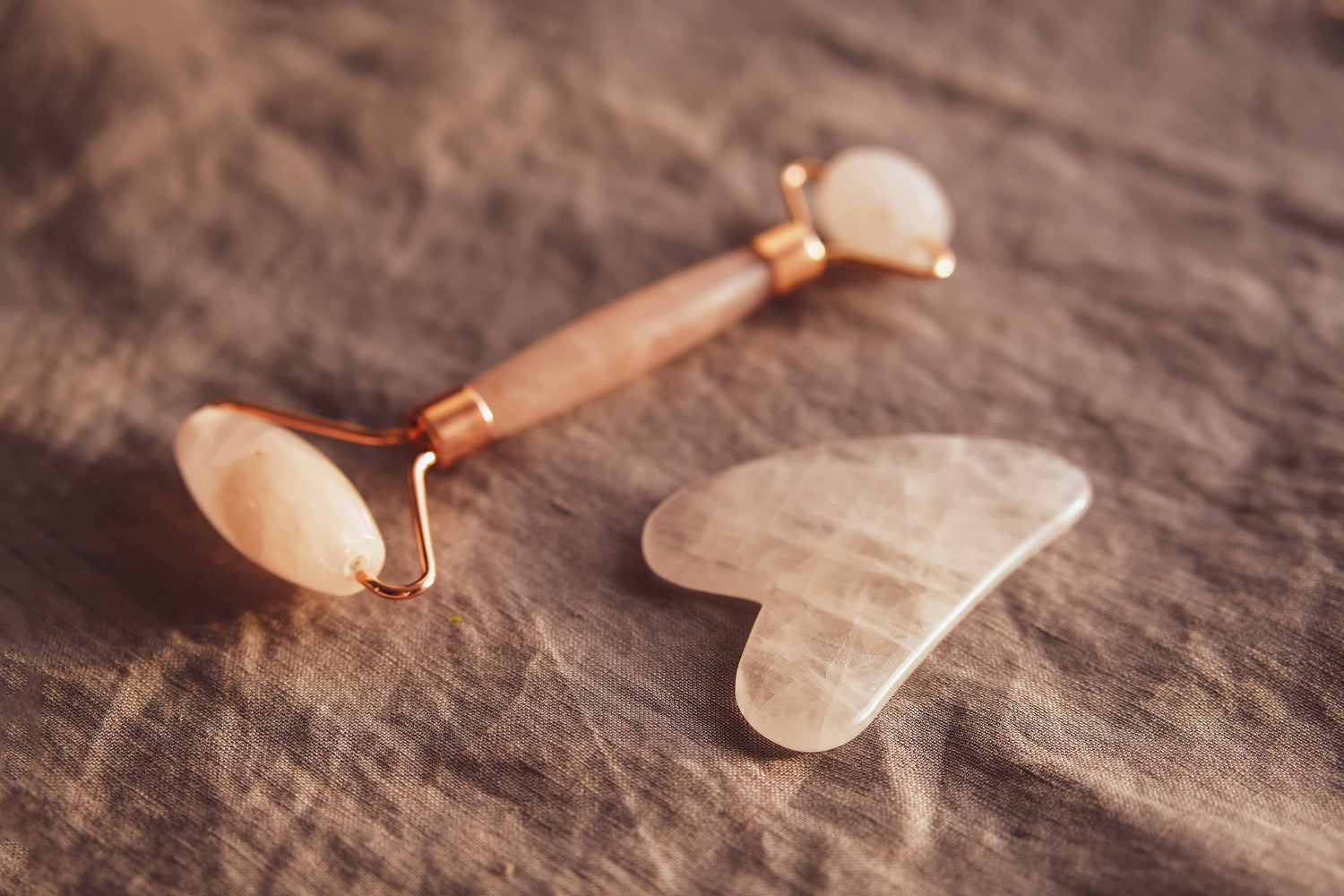 Un rouleau de massage pour le visage en quartz rose et un Gua Sha pour le visage en quartz rose sont posés sur un linge en lin de couleur brun terracotta.