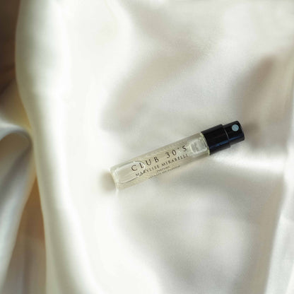 Un flacon de parfum en format 2ml du parfum Club 30's est posé sur un tissu en satin blanc.