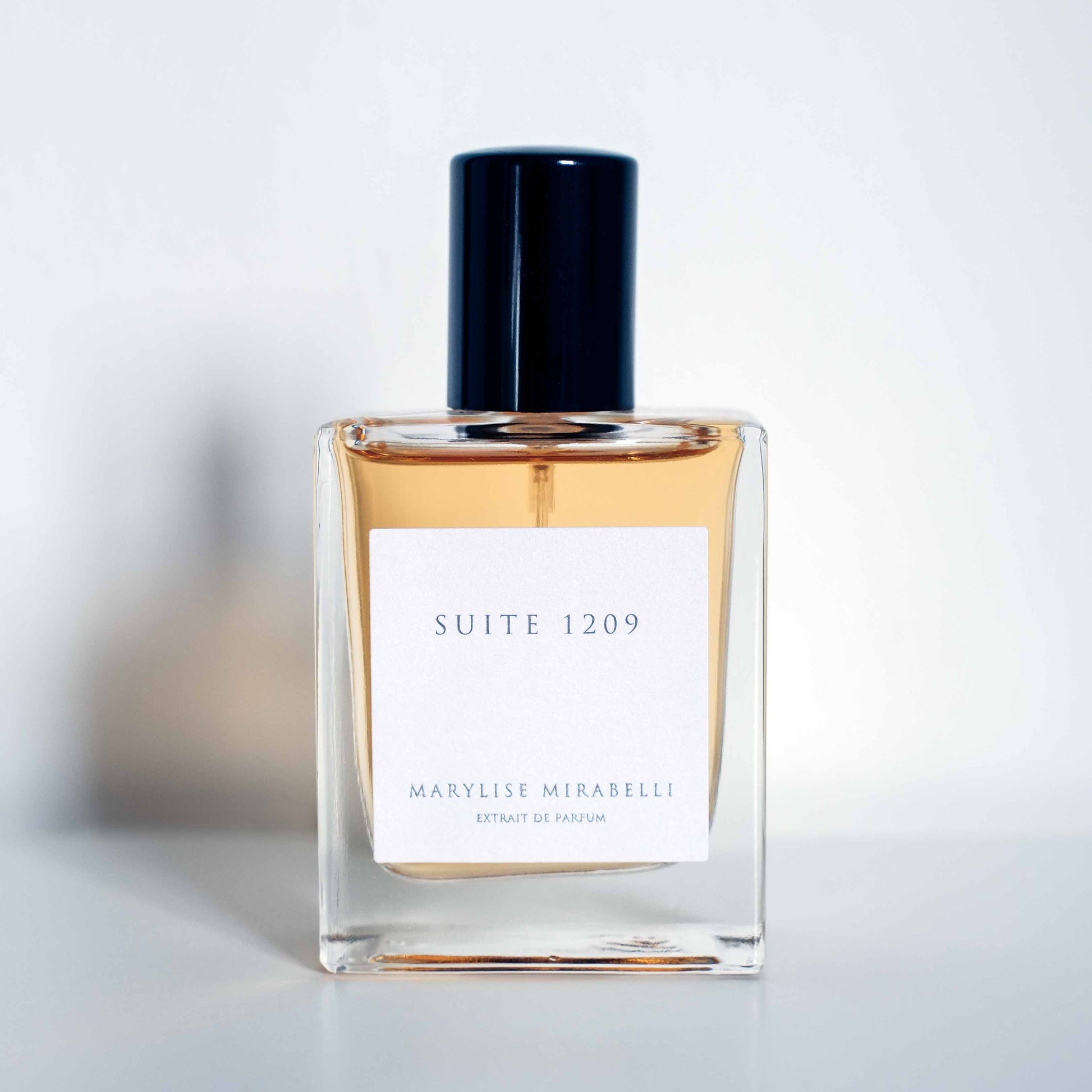 Flacon sobre et rectangulaire avec étiquette blanche et bouchon noir brillant, posé sur un fond blanc. Le jus du flacon est de couleur ambré. Sur l'étiquette il est inscrit Suite 1209 en haut et Marylise Mirabelli Extrait de parfum en bas.