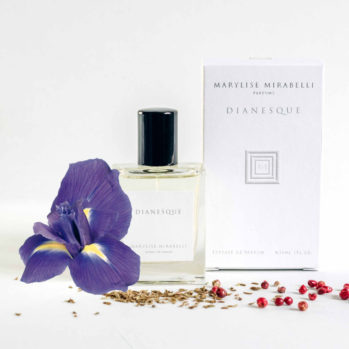 Petit flacon de parfum posé sur une table blanche à côté de sa boite. Le flacon de 30ml est rectangulaire avec une étiquette blanche, l'inscription Dianesque et un bouchon noir brillant. La boite est blanche avec le logo carré au centre et les inscriptions Marylise Mirabelli parfums et Dianesque en haut. Une fleur d'iris et posée sur le côté du flacon avec des baies roses et du cumin répandus sur la table.