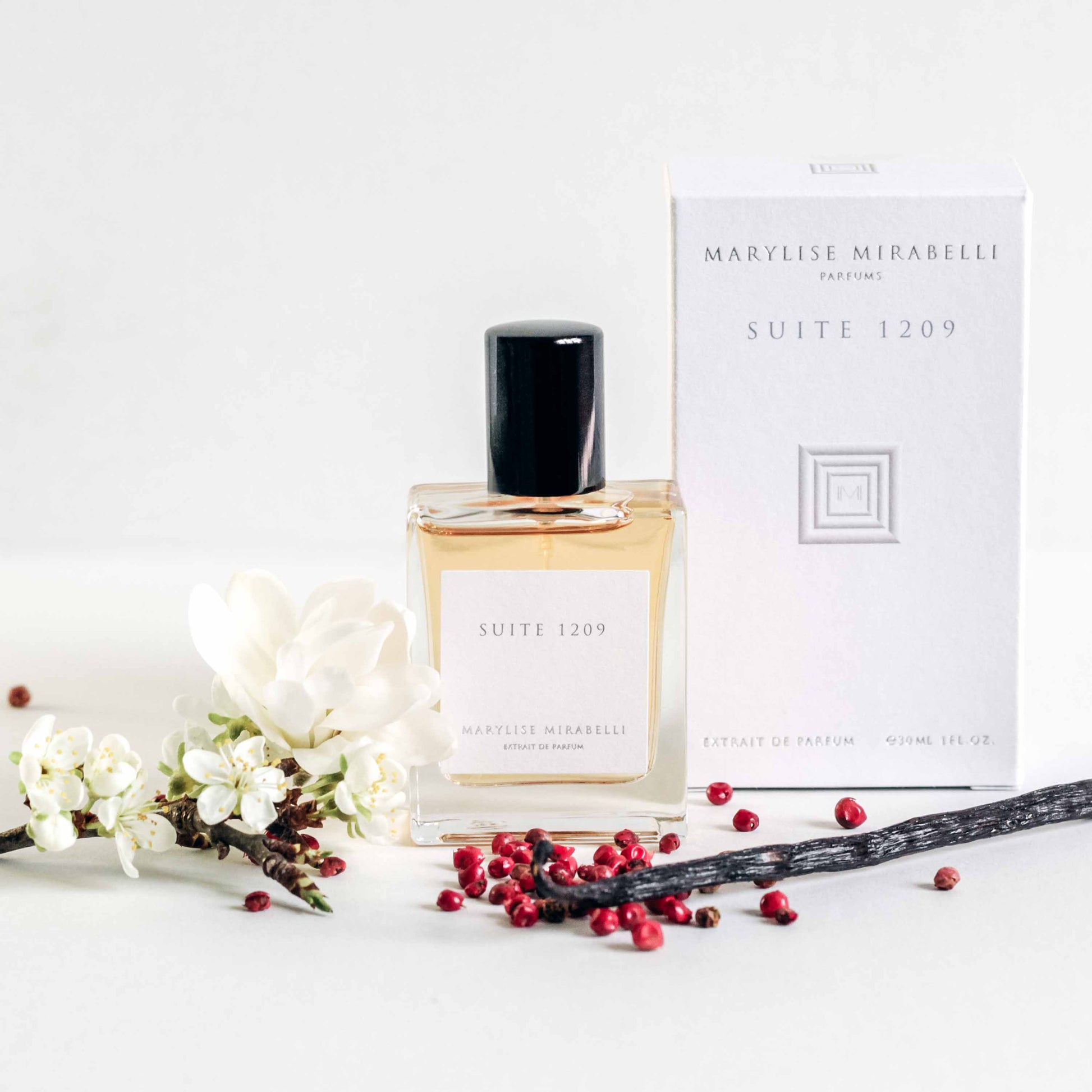 Petit flacon de parfum posé sur une table blanche à côté de sa boite. Le flacon de 30ml est rectangulaire avec une étiquette blanche, l'inscription Suite 1209 et un bouchon noir brillant. La boite est blanche avec le logo carré au centre et les inscriptions Marylise Mirabelli parfums et Dianesque en haut. Une petite branche de pommier avec de petites fleurs blanches et une fleur de camélia blanc son posés à gauche du flacon. Une gousse de vanille entourée de baies roses est posé de l'autre côté.