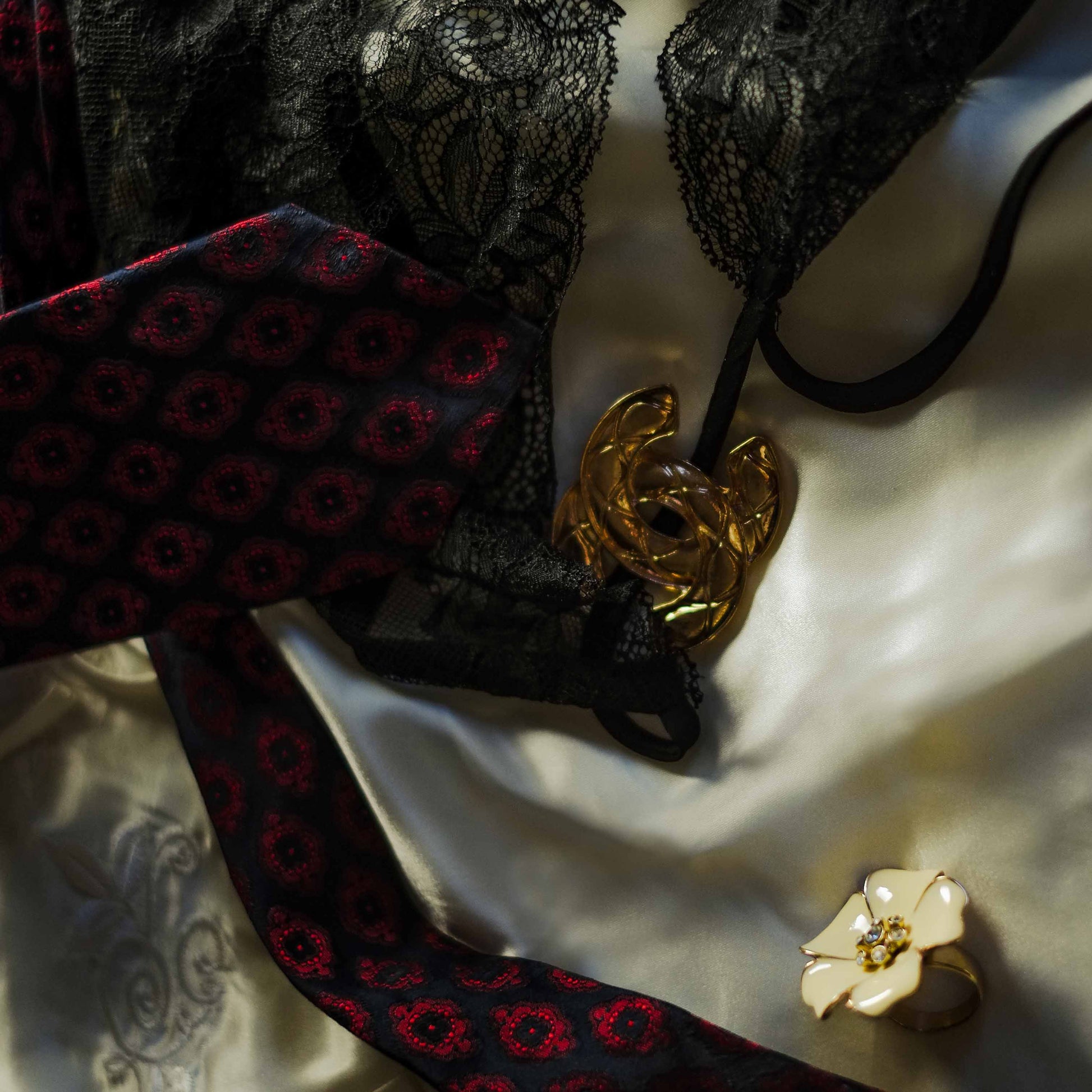 La photo représente l'univers chic et sensuel de la chanson "Who is It" de Michael Jackson. Une nuisette en dentelle noire, une cravate bleue nuit et rouge, une broche dorée Chanel et une bague en forme de fleur couleur crème sont posés sur un drap en satin crème.