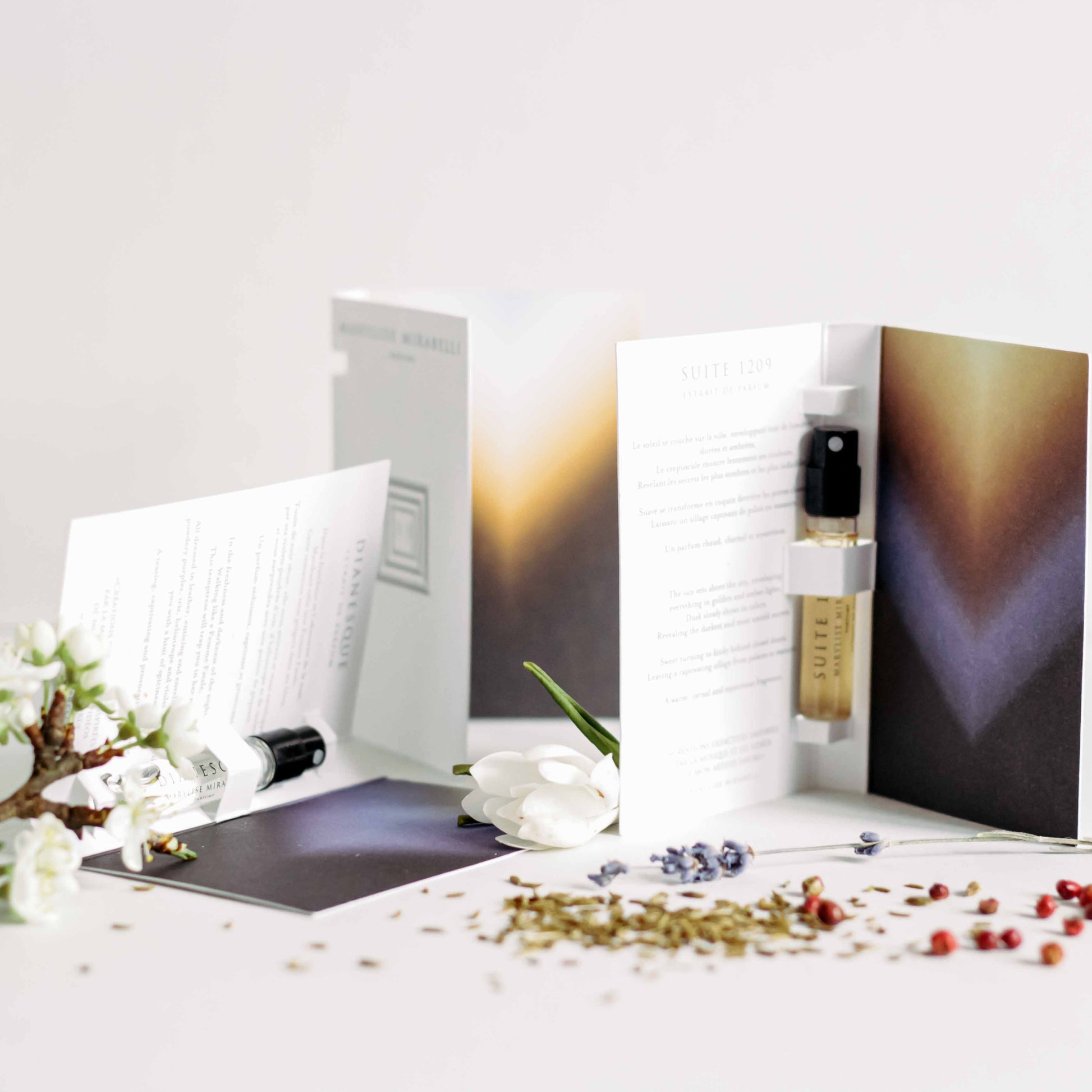 Le Set Découverte composé de 3 mini flacons en format 2ml accompagnés de leur cartonette descriptive. Les parfums sont disposés sur une table blanche et accompagnés d'une petite branche de pommier en fleur, d'une fleur de camélia blanc, d'une branche de lavande, de grains de cumin et de baies roses disposés sur la table.